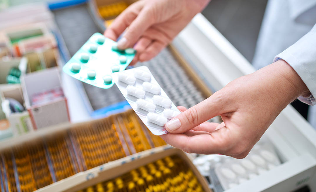 Two packs of pills being held above a drawer containing more pharmaceuticals